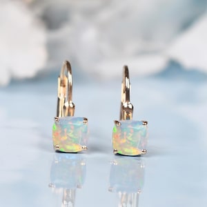 Peut inclure: Une paire de boucles d'oreilles à levier dorées avec des pierres d'opale carrées et iridescentes. Les opales présentent une gamme de couleurs, dont le blanc, le jaune et le vert. Les boucles d'oreilles sont posées sur un fond bleu clair.