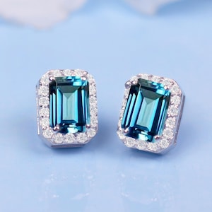 May include: A pair of silver earrings featuring a blue gemstone set in a halo of smaller diamonds.
