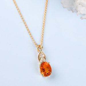 Pode incluir: Um colar dourado com um pingente laranja em forma oval. O pingente é adornado com pequenas pedras preciosas transparentes. O colar é exibido em uma superfície azul clara, com uma flor branca ao fundo.