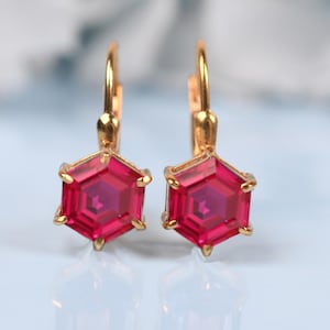 May include: A pair of gold-colored leverback earrings with hexagonal, ruby-red gemstones. The gemstones are faceted, catching the light and reflecting a deep, vibrant color. The earrings are displayed on a light blue surface, with a soft, blurred background.