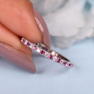 Peut inclure: Une bague délicate avec des pierres précieuses roses et claires alternées. La bague est tenue entre les doigts sur un fond bleu clair avec une fleur blanche floue. Les pierres précieuses sont serties dans une fine bande.