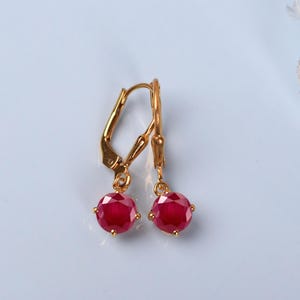 May include: A pair of gold-tone leverback earrings featuring round, faceted, deep red stones. The earrings have a polished gold finish and a secure hinged closure mechanism, showcasing a classic drop design with vibrant, saturated red gemstone accents.