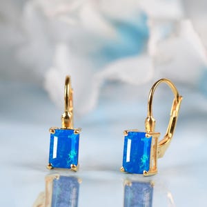 May include: A pair of gold-colored leverback earrings, each featuring a rectangular, vibrant blue gemstone. The earrings are set against a soft, blurred background, highlighting the jewelry's design and color.