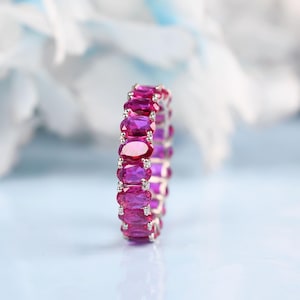 Puede incluir: Un anillo de plata con una banda continua de piedras preciosas rosas brillantes de talla ovalada. Las piedras están engastadas muy juntas, creando un diseño de anillo de eternidad completo. El anillo se muestra sobre un fondo suave y borroso.