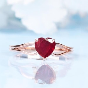 May include: A rose gold ring featuring a heart-shaped, deep red gemstone. The ring's band splits into two delicate strands on each side of the gemstone. The ring is set against a soft, light blue background, with a reflection visible below.