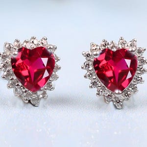 May include: A pair of heart-shaped ruby earrings with a halo of smaller clear stones. The earrings are silver.