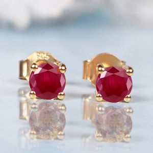 Op de afbeelding: Een paar gouden oorstekers, elk met een levendige, ronde, dieprode edelsteen. De oorbellen zijn gezet in een klassieke vier-klauwzetting, die licht reflecteert. De studs worden gepresenteerd op een reflecterend oppervlak.