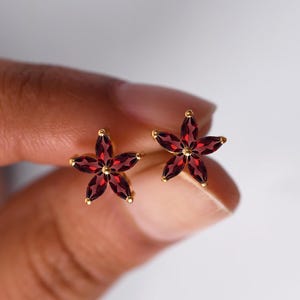 Könnte beinhalten: Ein Paar goldfarbene Ohrstecker, jeweils in Form einer fünfblättrigen Blume. Die Blütenblätter sind tiefrot, und die Mitte jeder Blume ist ein goldfarbener Akzent. Die Ohrringe werden in einer Hand gehalten.