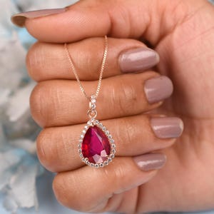 May include: A rose gold pendant necklace featuring a pear-shaped, faceted, deep red gemstone, surrounded by a halo of small, clear gemstones. The necklace is held against a soft background.