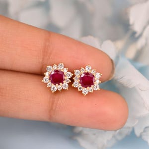 May include: A pair of gold stud earrings featuring a central round-cut deep red ruby gemstone surrounded by a halo of twelve small, clear, round-cut white diamonds. The earrings are designed in a floral shape with a polished gold metal setting.