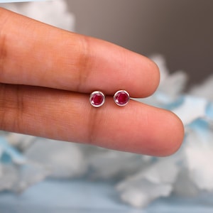 以下が含まれることがあります： 丸く鮮やかな赤い宝石を銀でセットした、小さなスタッドピアス。ピアスは人の指に表示され、そのサイズとデザインを示しています。これらのイヤリングは、クラシックなジュエリーアイテムです。