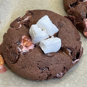 May include: A chocolate chip cookie with melted chocolate and marshmallows on top. The cookie is on a piece of parchment paper.