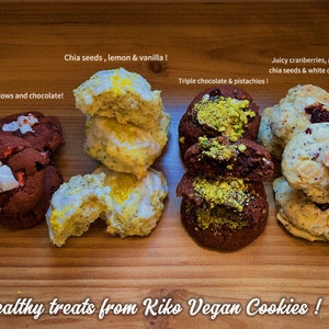 May include: Four different types of vegan cookies on a wooden surface. The cookies are arranged in four rows. The first row is a red cookie with white marshmallows and chocolate chips. The second row is a yellow cookie with lemon and vanilla. The third row is a brown cookie with chocolate and pistachios. The fourth row is a white cookie with cranberries, almonds, chia seeds, and white chocolate. The text "Healthy treats from Kiko Vegan Cookies!" is written below the cookies.