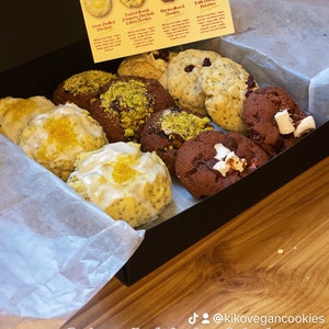 May include: A box of assorted vegan and gluten-free cookies. The cookies are arranged in a black box with white tissue paper. The cookies include lemon vanilla, toasted almond cranberry oat crumble,  marvellous chocolate, and triple chocolate & potato. The box has a sticker that says "Indulge yourself with a box of vegan & gluten free goodies!" and "Kiko Vegan Cookies".