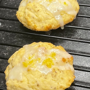 May include: Two golden brown, round, glazed cookies with a white glaze and yellow lemon zest on top. The cookies are on a black wire rack.
