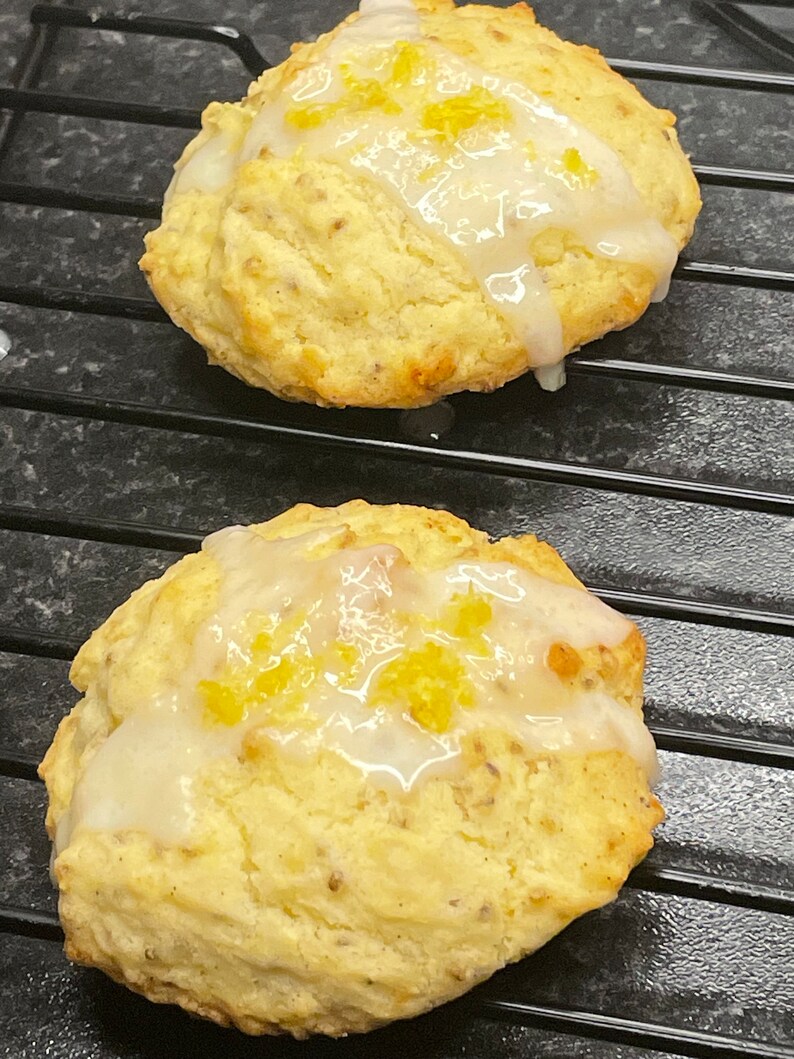 May include: Two golden brown, round, glazed cookies with a white glaze and yellow lemon zest on top. The cookies are on a black wire rack.