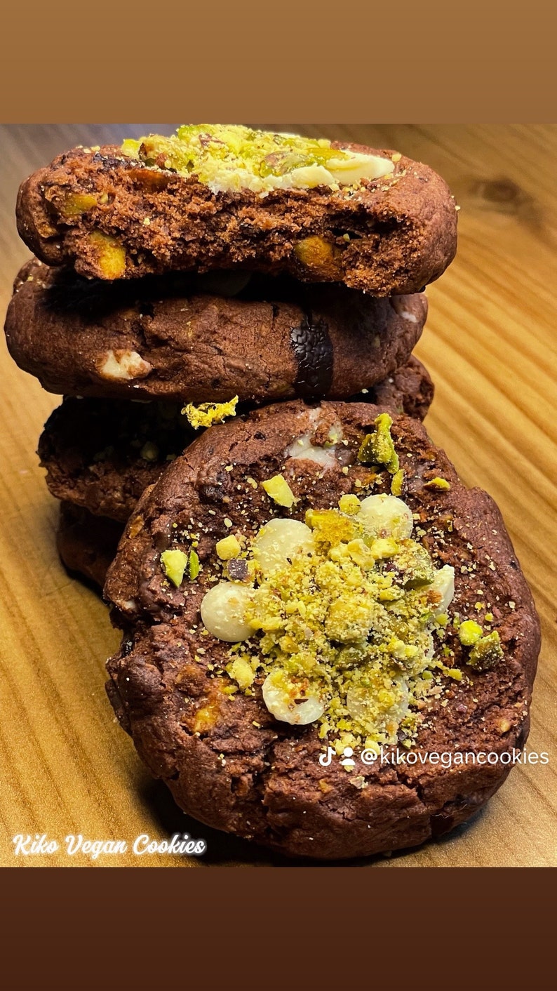 May include: A stack of four chocolate chip cookies topped with white chocolate chips and chopped pistachios. The cookies are on a wooden surface. The text "Kiko Vegan Cookies" is visible at the bottom of the image. The text "@kikovegancookies" is visible on the cookie.