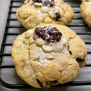 May include: Close-up of a white chocolate chip cookie with a cranberry on top. The cookie is dusted with powdered sugar and is sitting on a black wire rack.