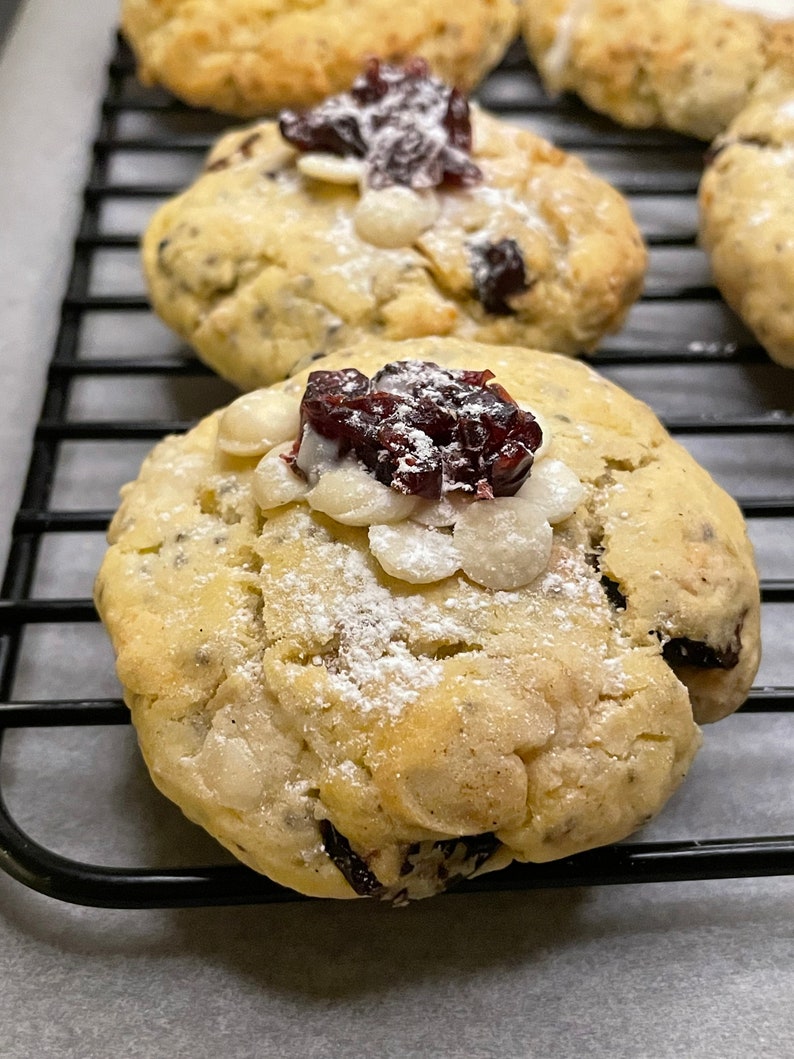 May include: Close-up of a white chocolate chip cookie with a cranberry on top. The cookie is dusted with powdered sugar and is sitting on a black wire rack.