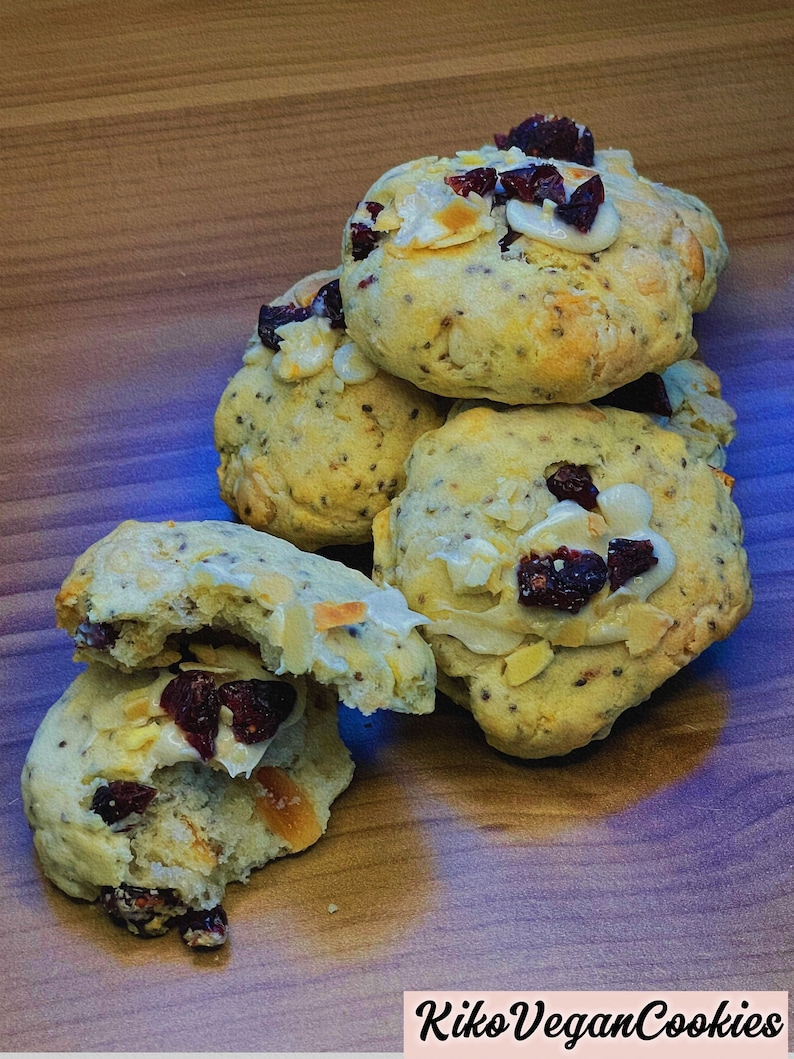 May include: A stack of four vegan cookies with white chocolate chips, cranberries, and a light glaze. The cookies are on a wooden surface. The text "Kiko Vegan Cookies" is visible in the bottom right corner of the image.