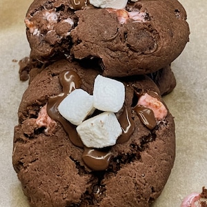 May include: Two chocolate chip cookies topped with mini marshmallows and drizzled with chocolate. The cookies are on a brown paper surface. The text "Kiko Vegan Cookies!" is written in pink letters at the bottom of the image.