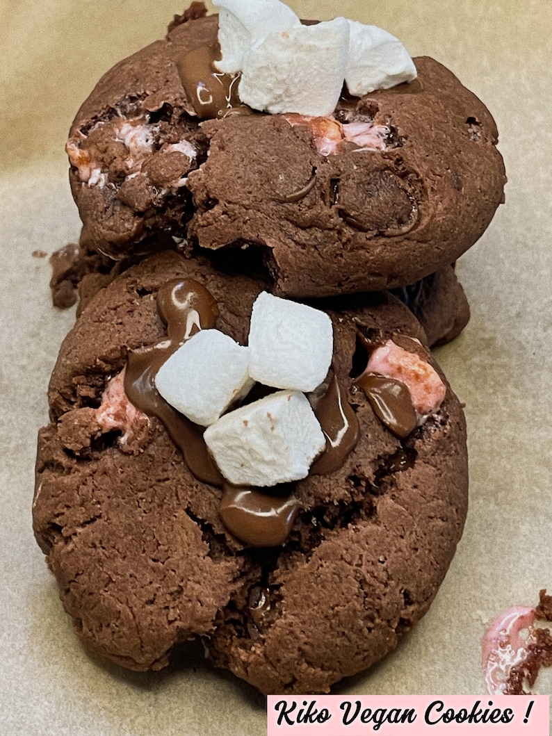 May include: Two chocolate chip cookies topped with mini marshmallows and drizzled with chocolate. The cookies are on a brown paper surface. The text "Kiko Vegan Cookies!" is written in pink letters at the bottom of the image.