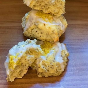 May include: Three vegan lemon poppyseed cookies with a white glaze and lemon zest on top. The cookies are stacked on top of each other on a wooden surface. The text "Kiko Vegan Cookies!" is written below the cookies.