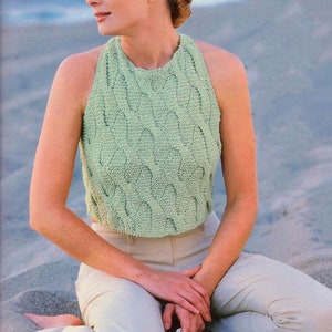 Puede incluir: Top halter verde claro con diseño tejido. El top se combina con pantalones claros. La imagen muestra a una persona sentada en arena, con sandalias blancas. El top es sin mangas y tiene un cuello alto.