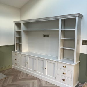 May include: White built-in entertainment unit with shelves and cabinets. The unit has a white painted finish and brass hardware. The unit is installed in a room with a herringbone patterned floor.