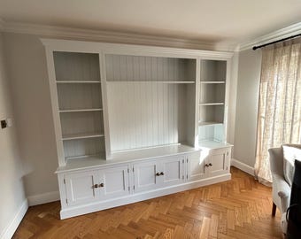 May include: White built-in bookcase with a white paneled back and a lower cabinet with doors. The bookcase has shelves and is in a room with a wood floor.