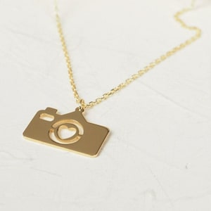 May include: A gold necklace with a camera pendant. The camera has a heart cut out in the center.