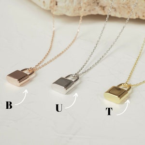 Peut inclure: Trois colliers avec pendentif en forme de cadenas dorés, un en or rose, un en argent et un en or. Les colliers sont sur un fond blanc avec un rocher derrière eux. Les pendentifs en forme de cadenas sont tous de la même taille et de la même forme.