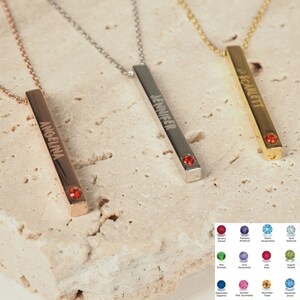 May include: Three gold bar necklaces with different names engraved on them: Angelina, Jennifer, and Scarlett. Each necklace has a small red gemstone at the end of the bar.