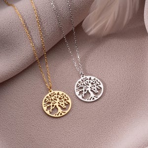May include: Two necklaces with tree of life pendants. One necklace is gold-colored, and the other is silver-colored. The pendants are circular with a detailed tree design. The chains are delicate, and the background is a soft, neutral fabric.