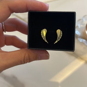 Puede incluir: Un par de pendientes de alas de &aacute;ngel de oro en una caja negra. Los pendientes son peque&ntilde;os y delicados.