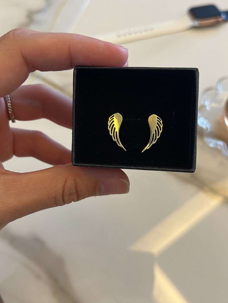 Puede incluir: Un par de pendientes de alas de &aacute;ngel de oro en una caja negra. Los pendientes son peque&ntilde;os y delicados.