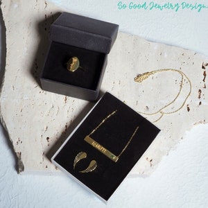 Puede incluir: Un conjunto de joyas de oro en cajas negras. El conjunto incluye un anillo con un dise&ntilde;o de flor, un collar con un colgante de barra que dice "SCARLETT" con una piedra verde, y un par de pendientes con alas de &aacute;ngel.