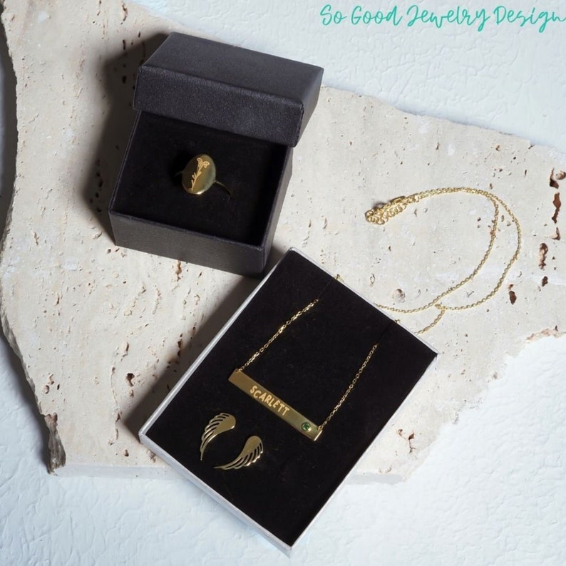 Puede incluir: Un conjunto de joyas de oro en cajas negras. El conjunto incluye un anillo con un dise&ntilde;o de flor, un collar con un colgante de barra que dice "SCARLETT" con una piedra verde, y un par de pendientes con alas de &aacute;ngel.