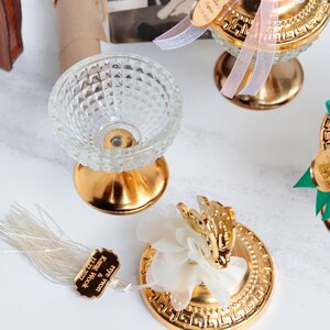 May include: Close-up of several decorative gold-colored pedestal bowls and decorative elements. One bowl is clear glass with a diamond pattern. Other elements include gold-colored bases, a butterfly, and a tag with text.