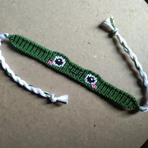 May include: Green friendship bracelet with white and black woven details, featuring two pink and black eyes.