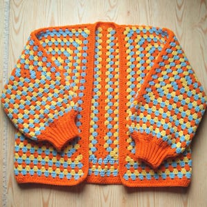 May include: A handmade, open-front crochet cardigan in a granny square pattern. The sweater features a vibrant color scheme of orange, yellow, and light blue squares, with solid orange trim and cuffs. The sleeves are long and the overall style is relaxed.