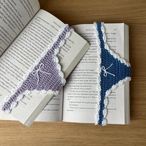 Könnte beinhalten: Zwei dreieckige Häkel-Lesezeichen, eines in Helllila und eines in Dunkelblau, jeweils mit weißem Bogenrand und einer kleinen weißen Schleife. Diese handgefertigten Textilaccessoires liegen in einem aufgeschlagenen Buch mit gedrucktem Text.