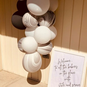 May include: A white sign with black text that reads "Welcome... to all the babies in the place with style and grace". The sign is leaning against a wall with a cluster of white and grey marble balloons.