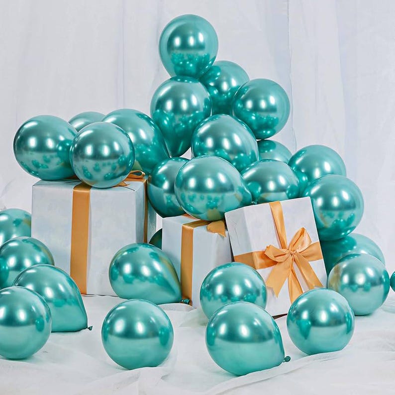 May include: A group of teal metallic balloons arranged in a pile on a white surface. The balloons are clustered together in a random pattern, with some balloons partially obscured by others. There are two white gift boxes with gold ribbon in the center of the image.
