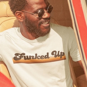 May include: A man wearing a white t-shirt with the text "Funked Up" in a retro font with brown and orange stripes. He is wearing sunglasses and sitting in a red car.