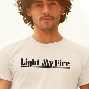 Op de afbeelding: Een wit T-shirt met korte mouwen en de tekst Light My Fire in het zwart op de borst gedrukt. De belettering is gestileerd in een vet, retro-geïnspireerd lettertype en wordt onderstreept door twee dunne horizontale zwarte lijnen.