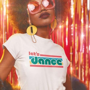 May include: White t-shirt with the text "let's dance" in a retro font. The shirt has a slim fit and is paired with red pants and a beige belt. The background features a shiny, colorful curtain.