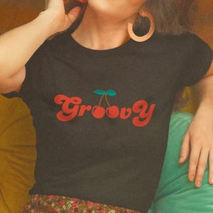 May include: A black short-sleeved t-shirt featuring the word Groovy in a red retro-style font, with two red cherries and a green stem replacing the double o in the text. The shirt has a crew neckline and a simple, fitted silhouette.
