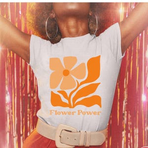 Op de afbeelding: Wit T-shirt met een oranje bloemenafbeelding en de tekst "Flower Power". Het shirt heeft een slanke pasvorm. De afbeelding toont ook een rode achtergrond en een beige riem.