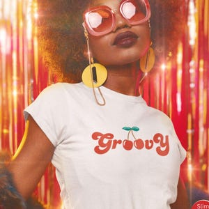 May include: A white short-sleeved t-shirt featuring the word Groovy in a red retro-style font with a small green and red cherry graphic above the letter v. The shirt has a slim fit cut and is displayed with large gold earrings and pink sunglasses.